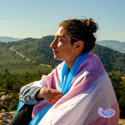 Young person wrapped in Transgender flag contemplating Transgender Day of Remembrance out in nature